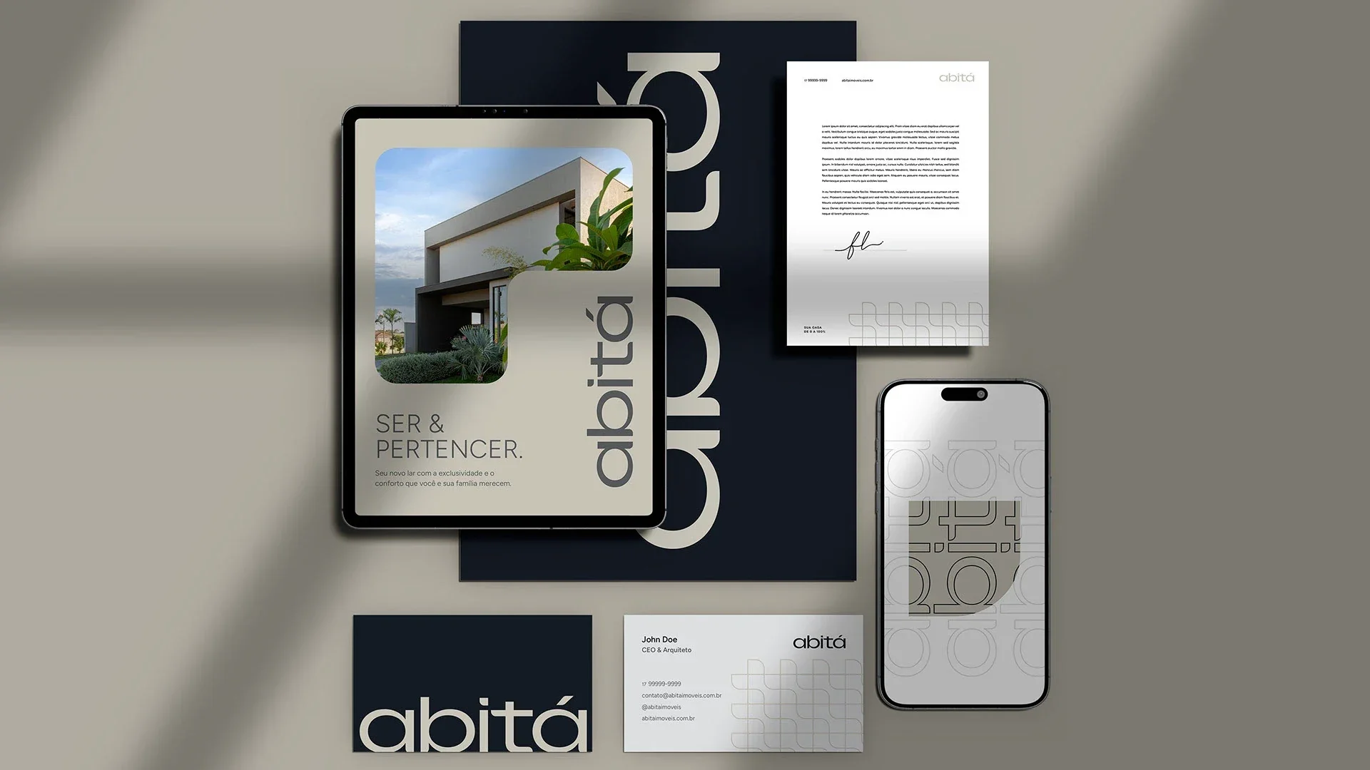 Composição da marca Abitá sob luz de veneziana: tablet com a peça “Ser & Pertencer”, papel timbrado, celular com o padrão do lettering, cartões e o nome em relevo sobre fundo bege.