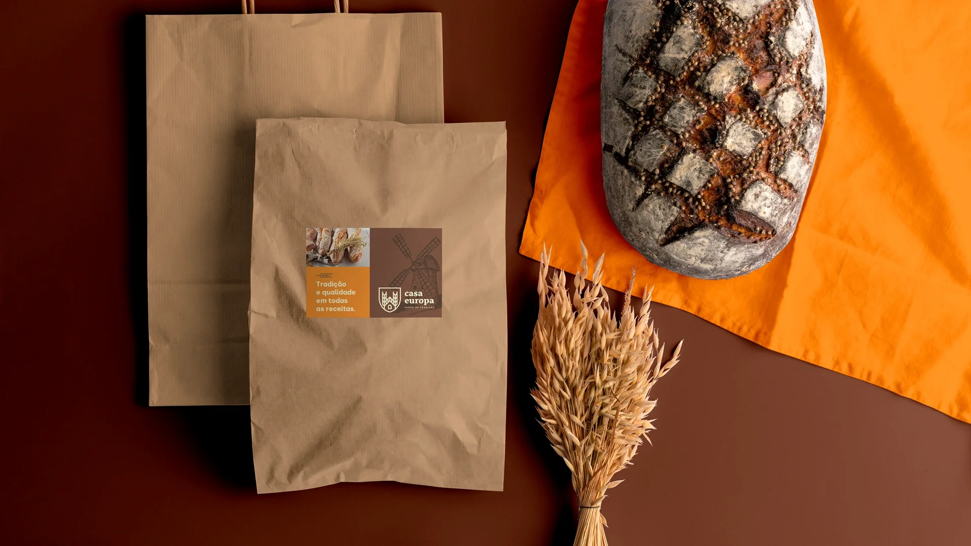 Duas sacolas de papel kraft da Casa Europa com a etiqueta “Tradição e qualidade em todas as receitas”, sobre fundo marrom, ao lado de um pão rústico sobre tecido laranja e um maço de espigas.