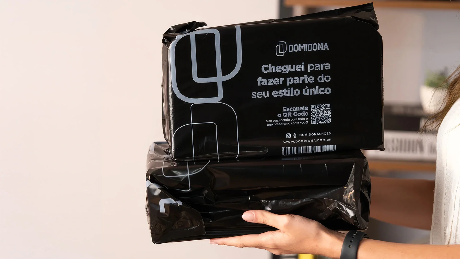 Duas embalagens pretas de e-commerce da Domidona nas mãos de uma cliente, com o símbolo em prata e a frase “Cheguei para fazer parte do seu estilo único”.