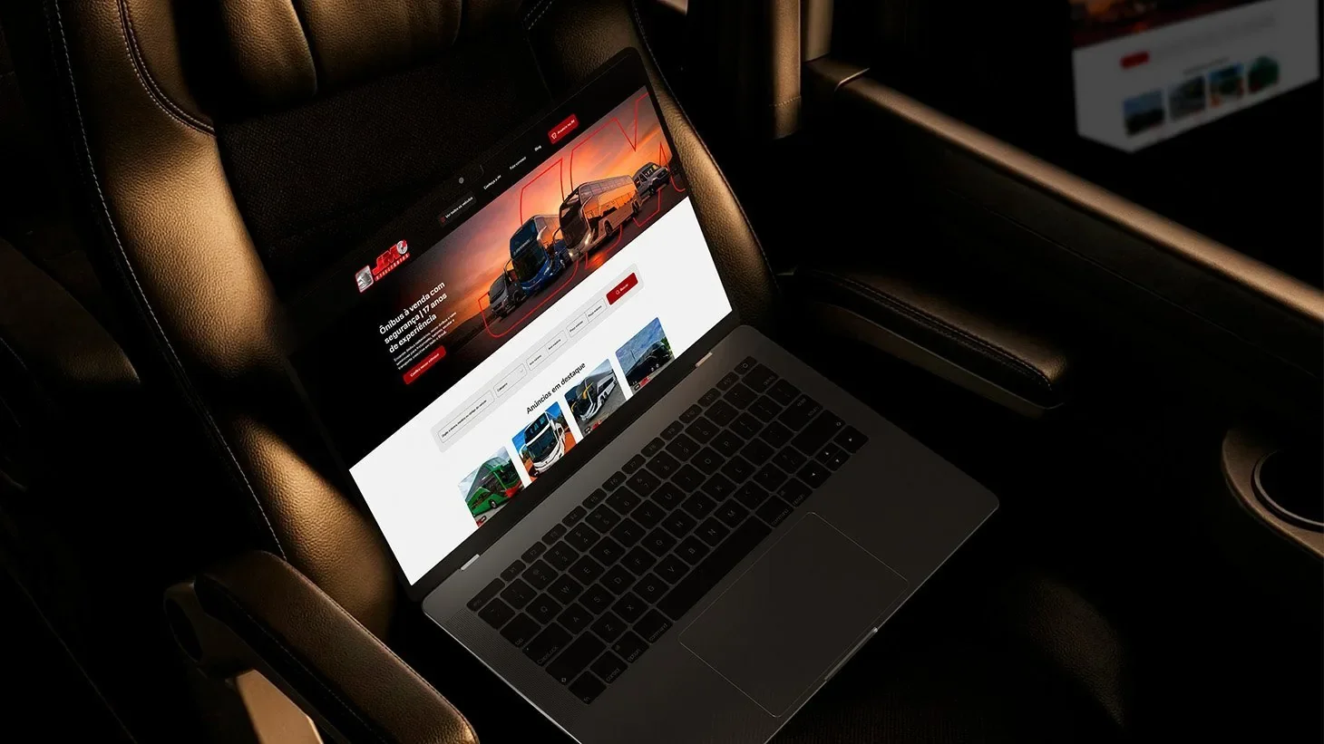 O site da JM Utilitários aberto num notebook, apoiado sobre o banco de couro de um ônibus, sob luz baixa.