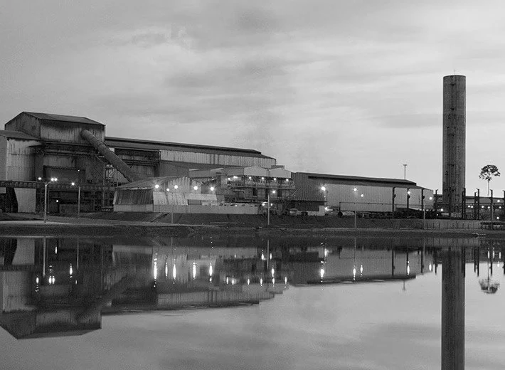 Foto em preto e branco da usina da SINOBRAS ao entardecer, com os galpões e a chaminé refletidos na água.