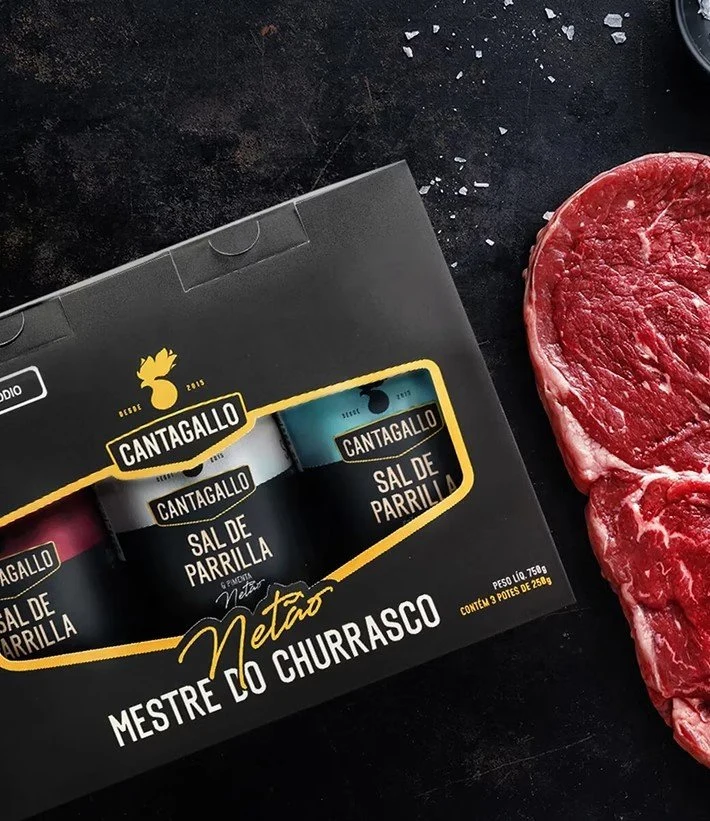 Kit Mestre do Churrasco da Cantagallo, com três potes de Sal de Parrilla na embalagem preta e dourada, ao lado de um bife cru sobre superfície escura.