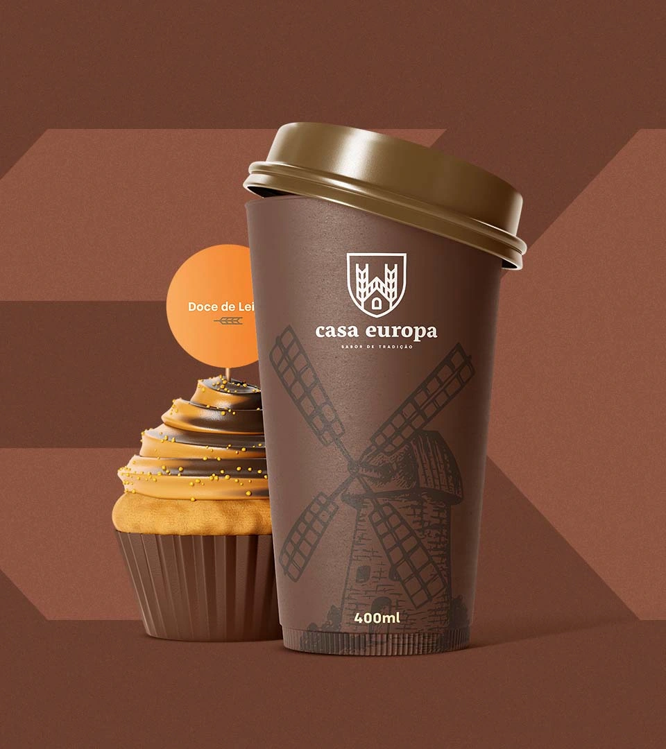 Copo de viagem de 400 ml da Casa Europa, em marrom com o moinho e a assinatura, ao lado de um cupcake de doce de leite com plaquinha laranja.