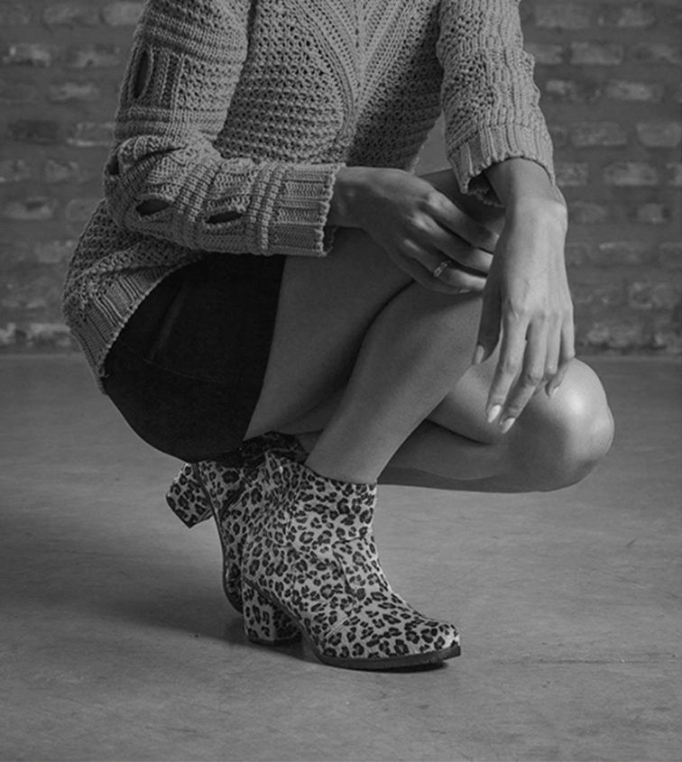 Foto em preto e branco: mulher agachada, de suéter de tricô e bota de oncinha, diante de uma parede de tijolos.
