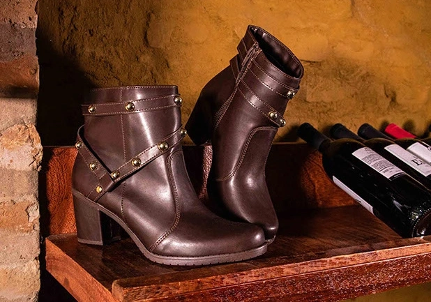 Bota marrom de cano curto com tachas douradas, apoiada em uma prateleira de madeira ao lado de garrafas de vinho.