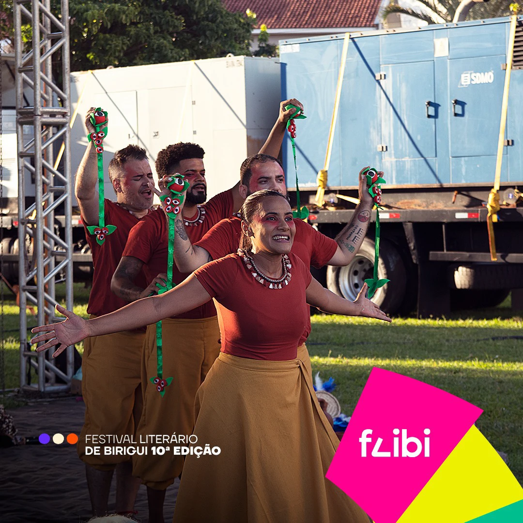 Grupo de dança em roupas vermelhas e ocre se apresenta ao ar livre, erguendo adereços verdes, com uma bailarina de braços abertos à frente.