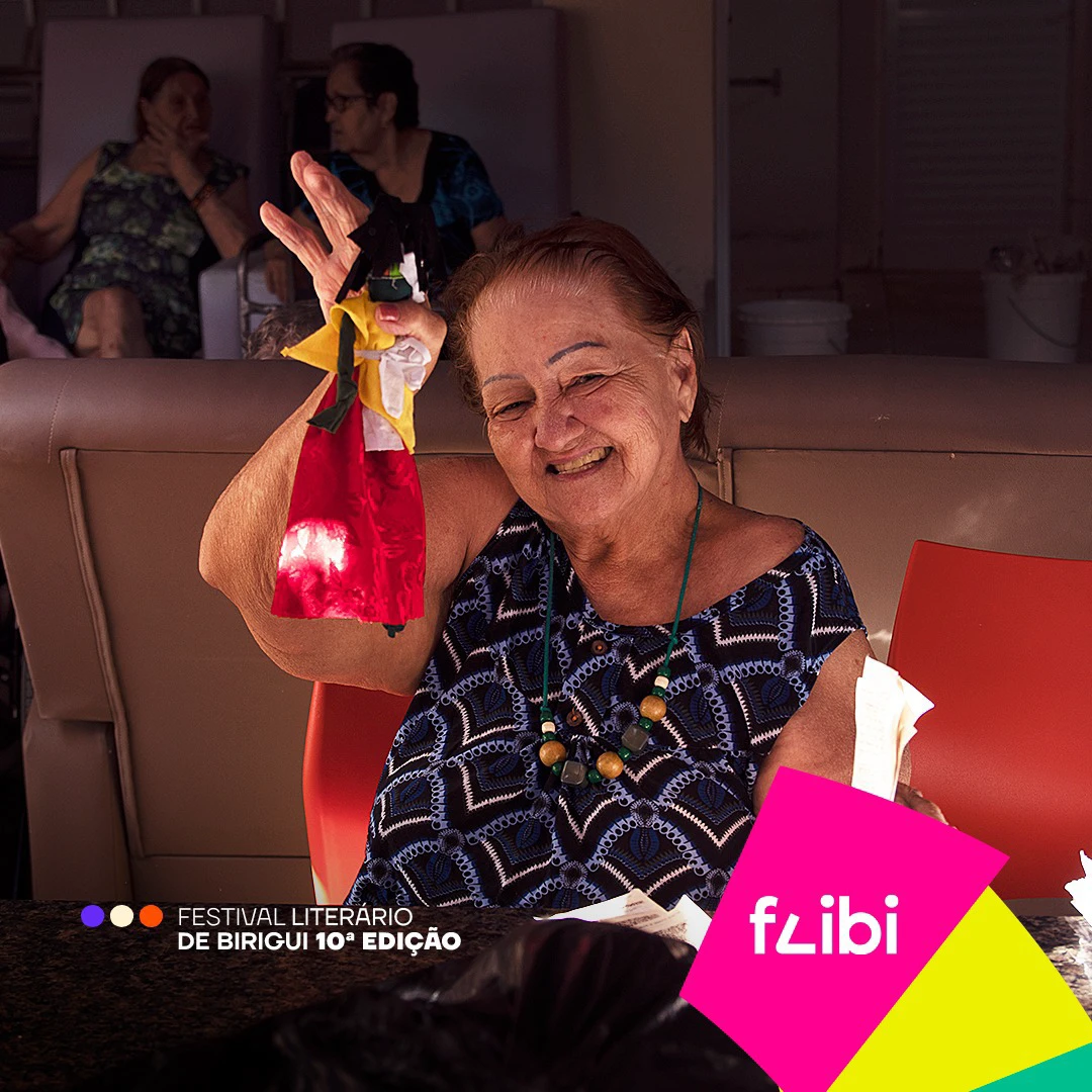 Senhora sorrindo enquanto ergue um fantoche de pano colorido, ao redor de uma mesa de atividades.