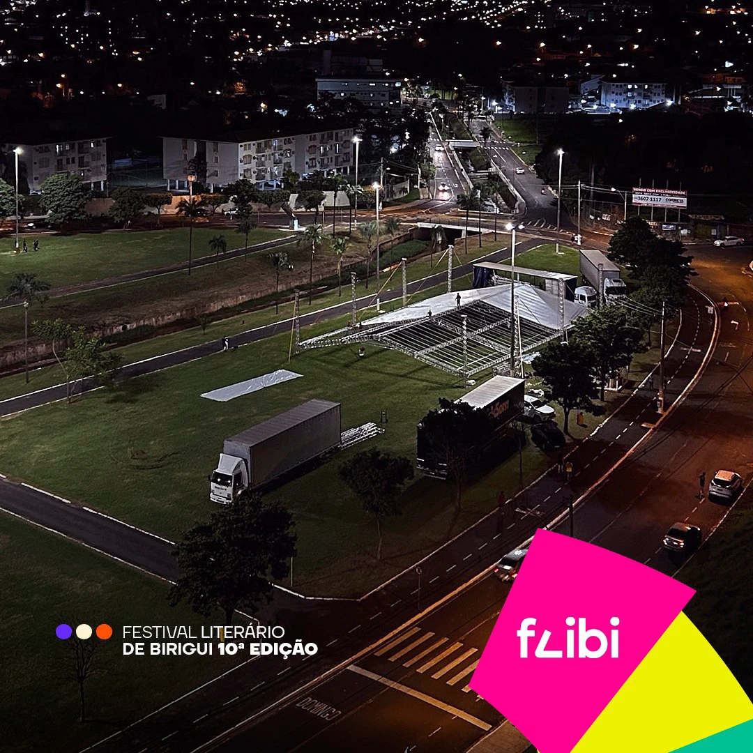 Vista aérea noturna do Parque do Povo durante a montagem do palco, com a estrutura metálica, caminhões e a cidade iluminada ao fundo.