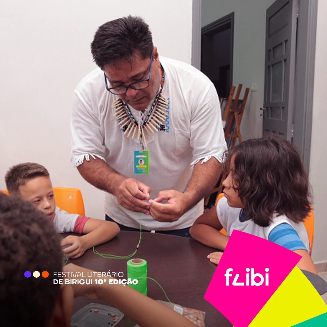 Oficina de artesanato com um educador indígena de colar de palha ensinando crianças a montar peças com linha verde.