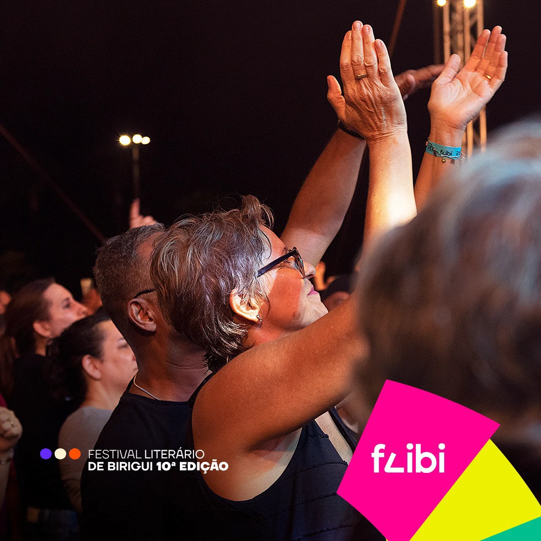Público do show à noite, com uma mulher de óculos aplaudindo de mãos erguidas e uma pulseira do Flibi no pulso ao lado.