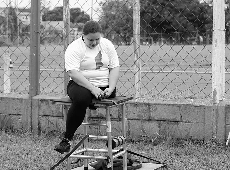 Atleta sentada num banco de treino, ao lado da prótese e dos equipamentos, numa pista ao ar livre.