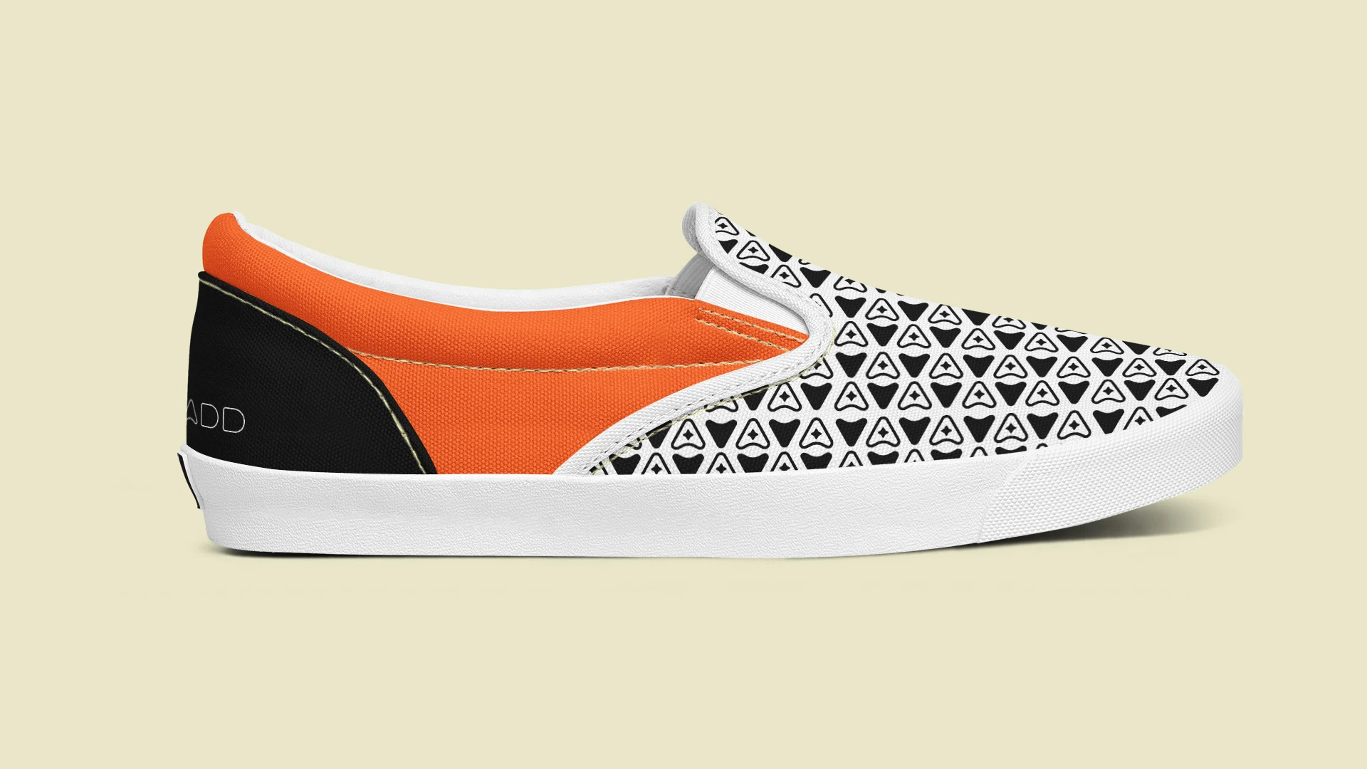 Tênis slip-on da Meadd de perfil sobre fundo bege, com o elástico laranja, o calcanhar preto com a marca em contorno e o cabedal branco padronizado.