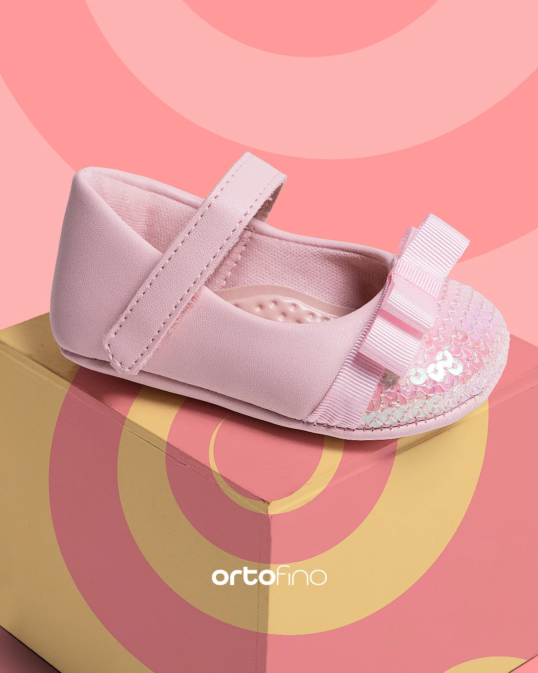 Post da Ortofino: sapatilha infantil rosa-claro com tira de velcro e laço, apoiada numa caixa amarela, sobre fundo rosa com arcos.