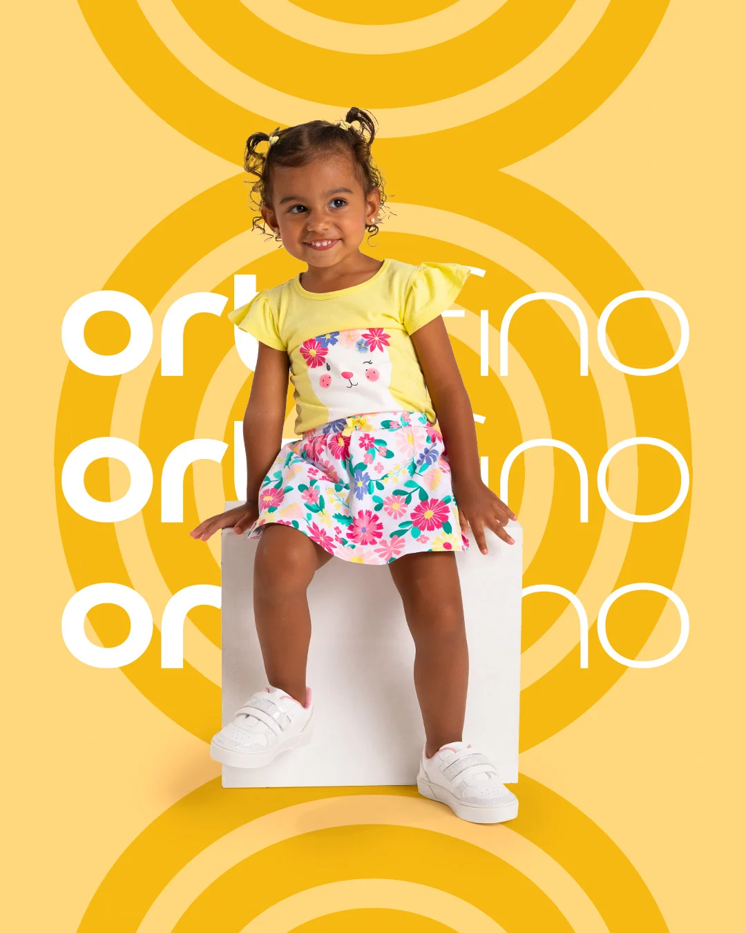 Post da Ortofino: menina de camiseta amarela e saia florida sentada num cubo branco, calçando tênis branco, sobre fundo amarelo com o logotipo repetido.