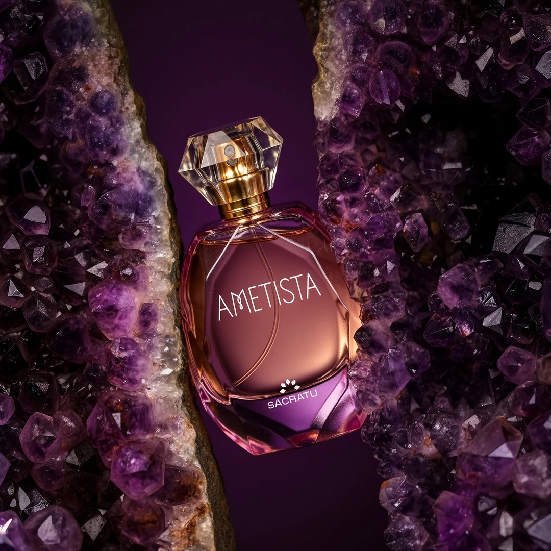Frasco do perfume Ametista, da Sacratu, com tampa facetada, apoiado na fenda de uma drusa de ametista roxa.
