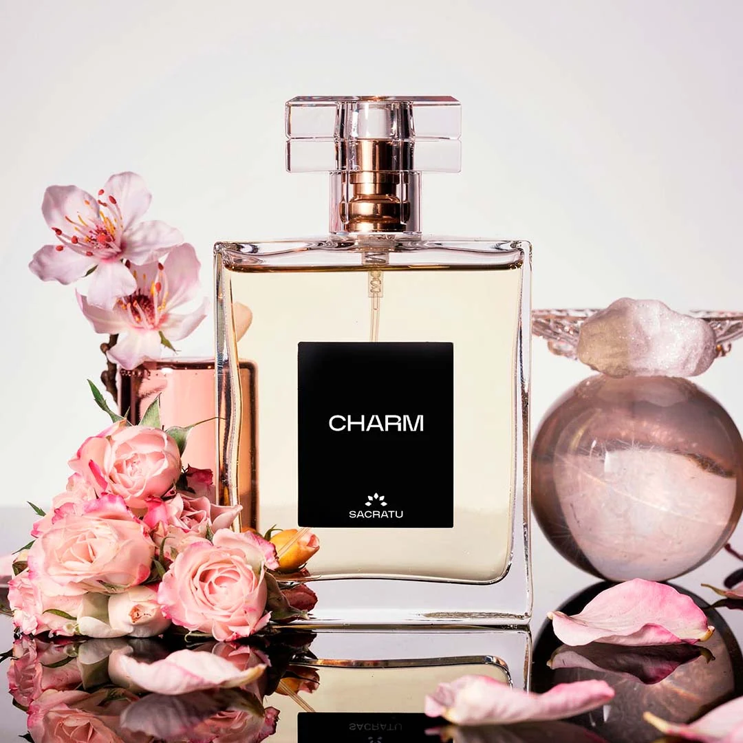Frasco do perfume Charm, da Sacratu, cercado de rosas e flores de cerejeira sobre fundo claro.