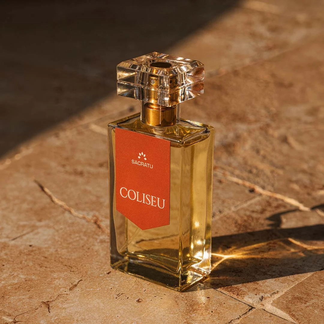 Frasco do perfume Coliseu, da Sacratu, de rótulo vermelho, sobre mármore travertino ao sol.