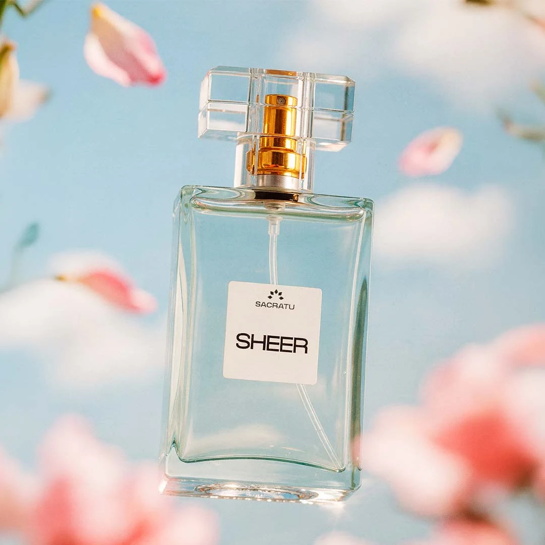 Frasco do perfume Sheer, da Sacratu, sobre fundo azul-claro com pétalas de cerejeira caindo.