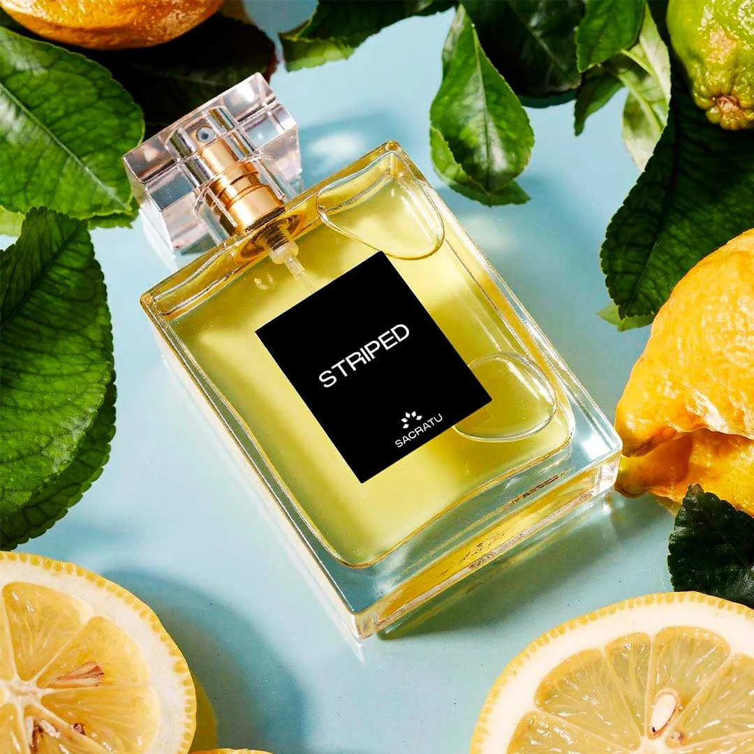 Frasco do perfume Striped, da Sacratu, entre folhas verdes e rodelas de limão sobre fundo azul-claro.